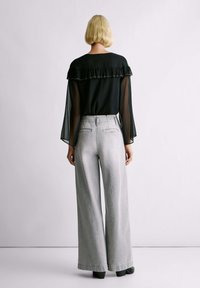 Osoba z krótkimi blond włosami, ubrana w przezroczystą czarną bluzkę z falbanami i szerokie jasnoszare spodnie, stoi na tle gładkiej jasnej ściany.