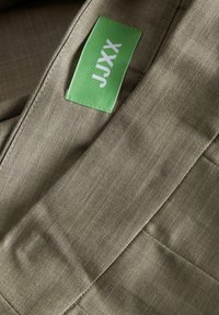 Tessuto beige con una sottile texture e cuciture diagonali; presenta un'etichetta verde con testo bianco che recita "JXXY."