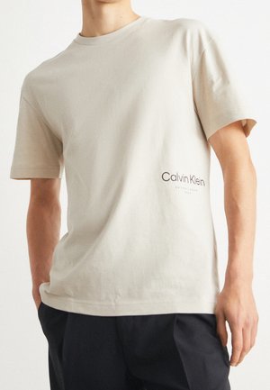 Osoba ubrana w jasnobeżową koszulkę Calvin Klein i ciemne spodnie, stojąca z jedną ręką w kieszeni na białym tle.