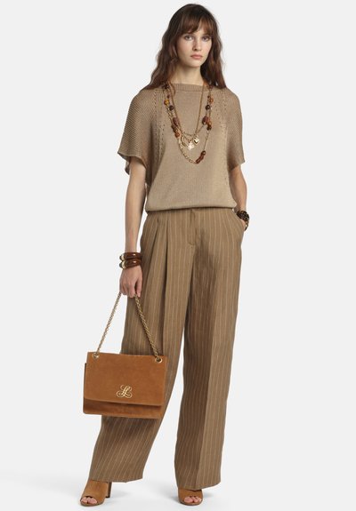 Femme portant un haut en maille beige avec des manches cape, un pantalon large à fines rayures marron, des colliers de perles superposés, des bracelets, tenant un sac à main en daim marron.