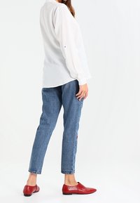 Blusa bianca a maniche lunghe con vestibilità ampia, jeans blu in denim alla caviglia e scarpe basse rosse, in piedi di spalle rispetto all'osservatore.