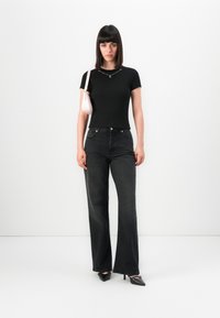 Woman with straight dark hair wearing black t-shirt, faded black jeans, black heels, silver necklace, and carrying a small white shoulder bag.