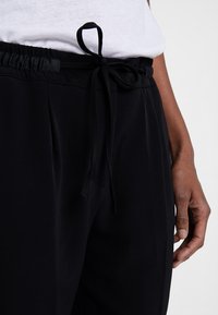 Pantalon de jogging noir texturé avec une taille à cordon de serrage. Comprend des poches latérales et des plis à l'avant, porté avec un haut blanc.