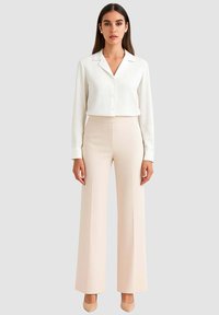 Donna in piedi che indossa una camicetta bianca con bottoni, pantaloni beige a vita alta e gamba larga, tacchi beige e orecchini di perle su uno sfondo grigio chiaro.