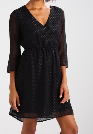Femme portant une robe noire à col en V avec des manches transparentes trois-quarts et un motif texturé à pois, debout devant un fond uni.