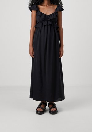 Kobieta w długiej czarnej sukience z marszczonymi ramionami i górą, w połączeniu z czarnymi masywnymi sandałami, stojąca na jednolitym tle.