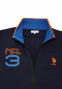 Námořnická bunda na zip s modrým a oranžovým límcem, "NR. 3" v oranžové a modré na levé straně hrudi a oranžové logo hráče polo USPA na pravé straně hrudi.