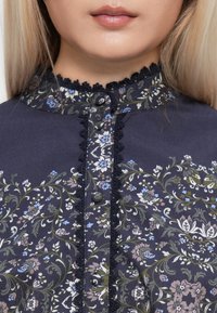 Close-up of a woman’s neck and chin wearing a navy floral blouse with lace trim and fabric-covered buttons at the collar and front.