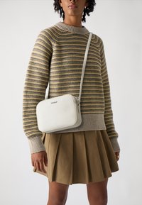Witte crossbody tas met een slank rechthoekig ontwerp, gladde lederen textuur en metallic logo, gecombineerd met een gestreepte trui en een geplooide rok.