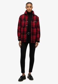 Red and black plaid jacket with a collar, two front pockets, button closures, worn over a black turtleneck, paired with black leggings and shoes.