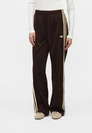 Άτομο που φοράει σκούρο καφέ παντελόνι Adidas με μπεζ ρίγες και μπεζ παπούτσια, στέκεται μπροστά σε απλό φωτεινό φόντο.