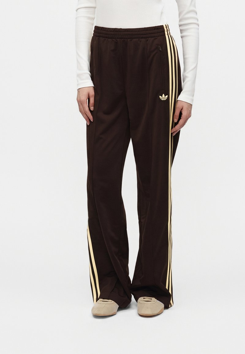 Άτομο που φοράει σκούρο καφέ παντελόνι Adidas με μπεζ ρίγες και μπεζ παπούτσια, στέκεται μπροστά σε απλό φωτεινό φόντο.