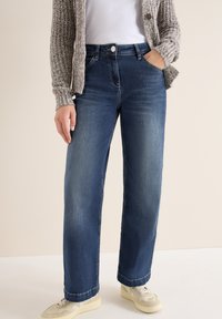 Vaqueros anchos azules con una textura suave, presentan un diseño clásico de cinco bolsillos y un cierre frontal con botón. Combinados con zapatillas blancas.