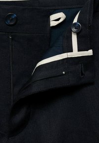 Pantaloni in tessuto blu scuro, con cerniera visibile, chiusura con bottone e bordo bianco sporco lungo l'interno. Superficie texturizzata con cuciture visibili.