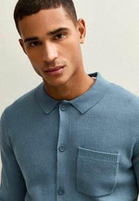 Ljusblå stickad polo med krage, som har en ficka med patch och knappstängning, texturerad tyg och en figursydd design.