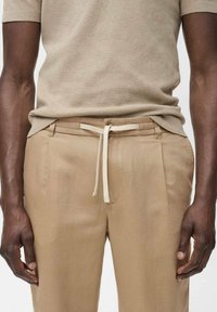 Pantalon en mélange de lin beige avec une taille à cordon, doté de poches latérales et d'une texture lisse. Associé à un haut en tricot beige assorti.