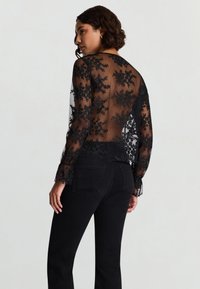 Camicia nera trasparente con ricamo floreale, maniche lunghe e apertura sulla schiena. Abbinata a jeans neri a vita alta.