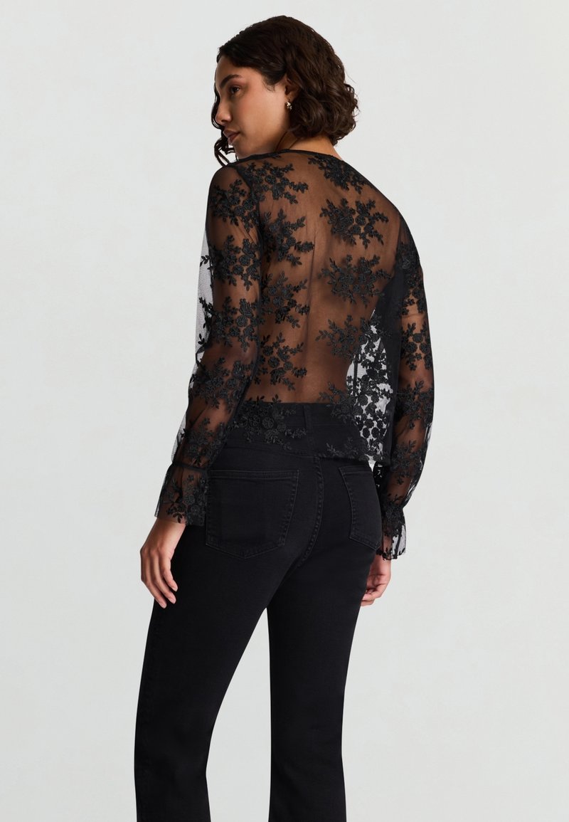 Camicia nera trasparente con ricamo floreale, maniche lunghe e apertura sulla schiena. Abbinata a jeans neri a vita alta.
