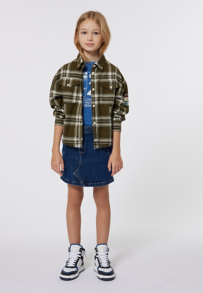 Groene geruite flanellen shirt met twee borstzakken, gedragen over een blauwe graphic t-shirt, gecombineerd met een spijkerrok en hoge sneakers.