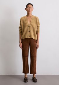 Beige gestrickte Strickjacke mit tiefem V-Ausschnitt, Knopfdetails, kombiniert mit braunen Leder-Breitabzugshosen und dunklen Slippern, vor einem neutralen Hintergrund.