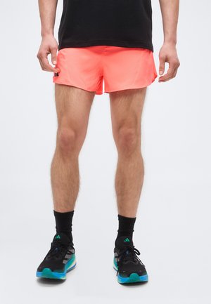 Persoană purtând pantaloni scurți sportivi coral strălucitor, șosete negre și pantofi de alergare negri cu accente turcoaz și albastre, stând pe fundal alb.