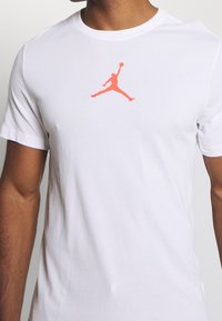 Vit bomulls-t-shirt med en ljusorange Jumpman-logotyp på bröstet, med rund hals och korta ärmar.
