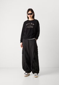 Svart hettegenser med sølv "DKNY Jeans 1989" logo, parret med løse svarte cargo-bukser med flere lommer og snøring i mansjettene. Sølv sneakers.