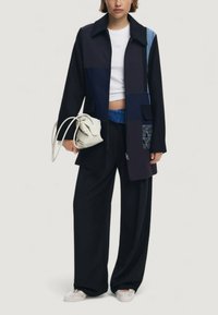Giacca patchwork nera con texture diverse, dettagli in denim, tasche con bottone, pantaloni neri larghi e scarpe chiare.