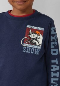 Felpa blu navy con texture a coste, con grafica di un rinoceronte in bicicletta. La manica sinistra presenta la scritta "WILD TAILS".