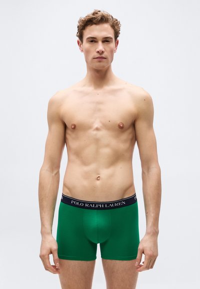 Νεαρός άνδρας με ανοιχτόχρωμο δέρμα και σγουρά μαλλιά, φορόντας πράσινα boxer briefs Polo Ralph Lauren, όρθιος μπροστά σε απλό ανοιχτό φόντο.