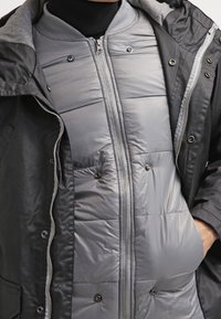 Personne portant une veste grise en couches et un manteau noir, avec une fermeture éclair et des boutons-pression visibles, la main dans la poche du côté droit.