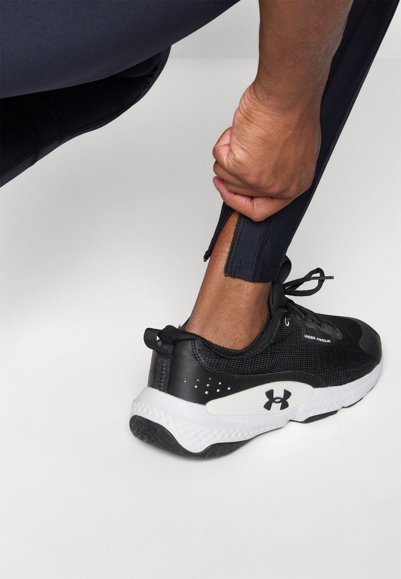 Zwarte sportieve schoen met een geweven bovenkant, witte middenzool en prominent logo. Zichtbare korte rits op de zoom van de marineblauwe broek.