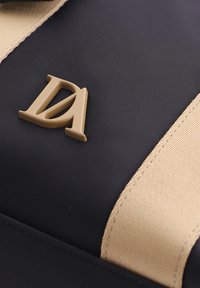 Logo DA beige sobre tela negra con correa de tela beige cosida a lo largo del lado, mostrando detalles de textura y material.