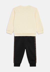 Kremowa bluza z ribbowymi mankietami, połączona z czarnymi spodniami dresowymi z brązowym paskiem z nadrukiem zwierzęcym po bokach. Miękki, codzienny materiał bawełniany.