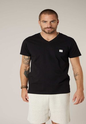 Homme aux cheveux courts et à la barbe portant un t-shirt noir à col en V et un short blanc, montrant des tatouages sur les deux bras et des bracelets au poignet gauche.