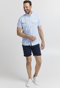 Camisa de manga corta azul claro con cierre de botones, que presenta detalles bordados y parches. Combinada con pantalones cortos azul marino y zapatillas blancas.