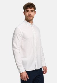 Homme aux cheveux courts et bouclés, portant une chemise blanche boutonnée et un pantalon sombre, debout devant un fond blanc uni.
