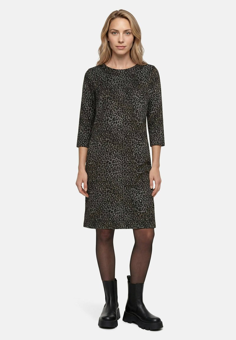 Femme debout portant une robe noire et grise imprimé léopard longueur genou, manches trois-quarts, collants noirs transparents et bottes noires épaisses.