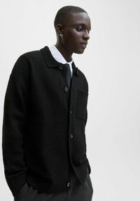 Melnas vilnas kardigāns ar apkakles dizainu, priekšējiem pogām, vienu krūšu kabatu un ribotu piekari. Uzvilkts virs baltas krekla un kaklasaites.