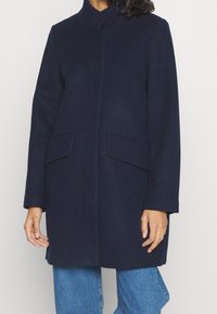 Marinblå ullrock med hög krage, sidofickor och rak skärning. Har en mjuk textur och minimalistisk design.