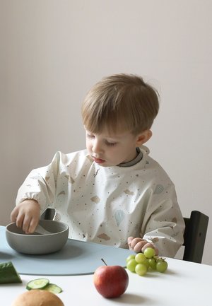 Malen otrok v vzorčastem podbradku sedi za mizo in meša hrano v skledi, v bližini pa so jabolko, grozdje, kumara in kruh.