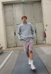 Hellblaues Kabelmuster-Sweatshirt, blassrosa Shorts, weiße Socken und pastellfarbene Sneakers. Geht über eine strukturierte Fläche in der Nähe einer grauen Tür.