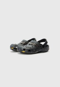 Black clogs made of rubber with ventilation holes, adjustable heel straps, and yellow accents on the sole. Textured surface and rounded toes.