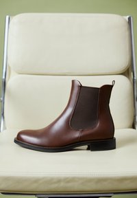 Brown leather Chelsea boot with elastic side panel and black sole placed on a cream-colored cushioned chair with metal frame.