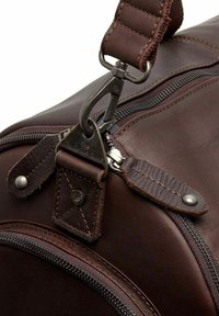 Primer plano de un bolso de viaje de cuero marrón, con un zipplate plateado, clip de metal y correa de cuero con detalles de costura. Los acentos incluyen una etiqueta de marca.