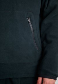 Mörkgrön fleecepullover med en dragkedjeficka och elastiska ärmslut. Mjuk textur och enfärgad design med platta sömmar.