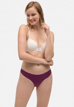 Sostén acolchado beige con tirantes ajustables, acompañado de un tanga morado con bordes en forma de scallop. La tela parece suave y lisa. El modelo posa de forma natural.