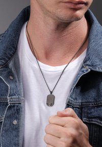 Zilveren dog tag ketting met een rechthoekige vorm, hangend aan een dunne ketting, gedragen over een wit t-shirt en onder een denim jas.