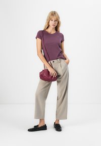 Frau in einem lila T-Shirt, beigen weiten Hosen, schwarzen Loafers und einer burgunderfarbenen Umhängetasche, die vor einem weißen Hintergrund steht.