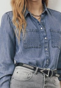 Camicia di denim con tasche sul petto decorate con borchie, bottoni a scatto argentati, collana a catena a più strati, jeans grigi chiari e cintura nera con fibbia triangolare argentata e motivo a freccia.